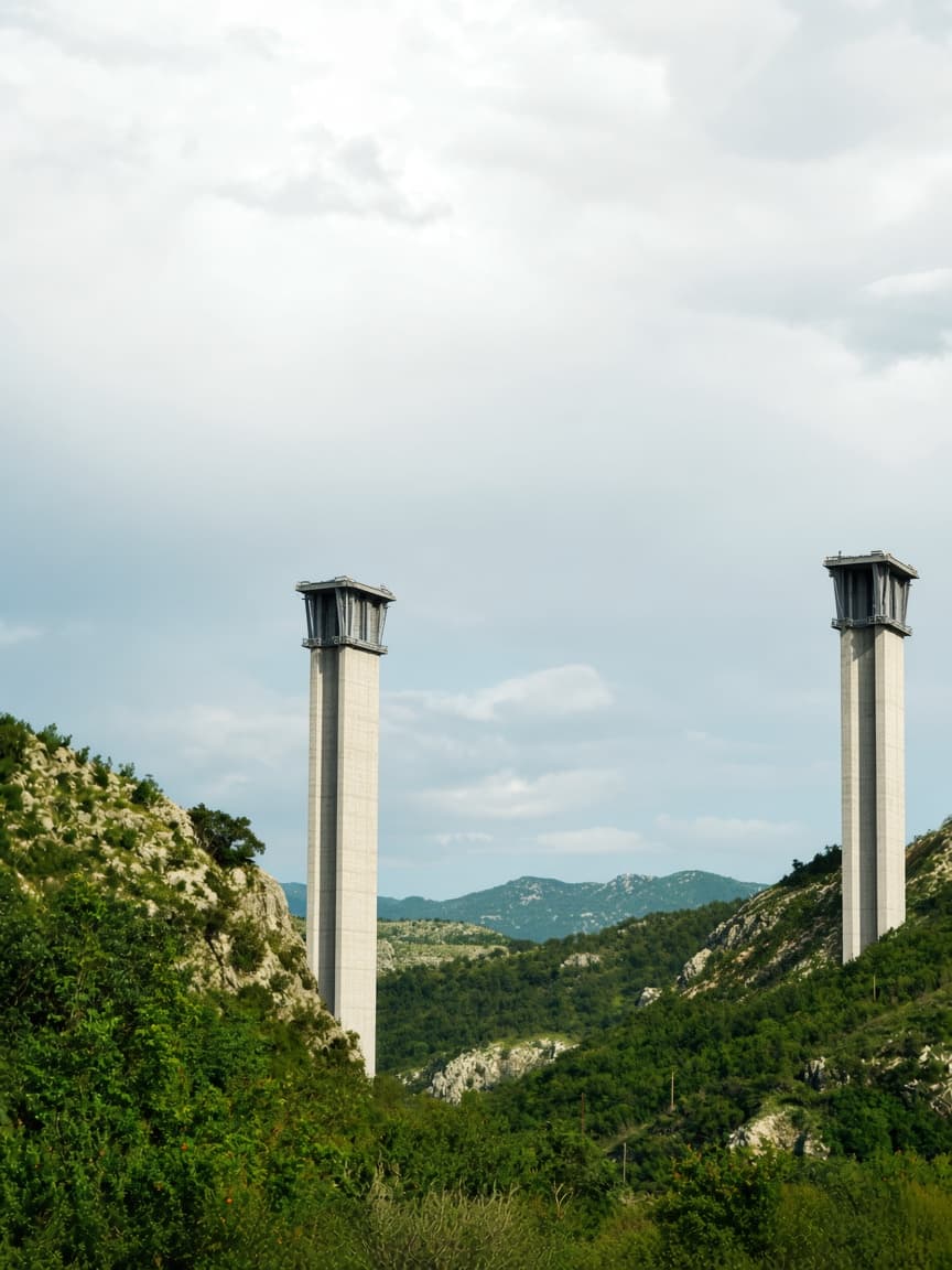 Two tall, slender concrete bridge pillars stand completed in a green, mountainous valley.