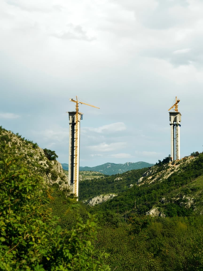Two tall concrete bridge pillars with cranes in a green, rocky mountain landscape.