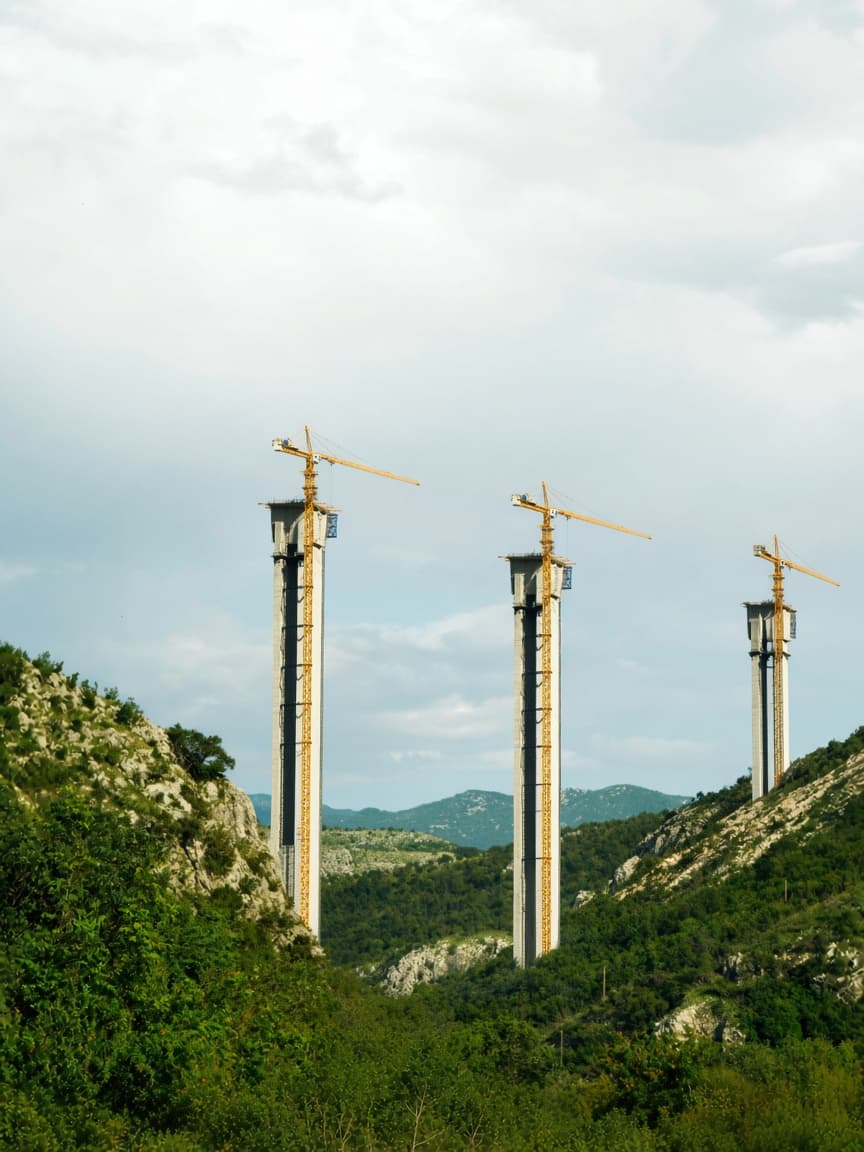 Three tall concrete bridge pillars under construction with cranes in a green valley.